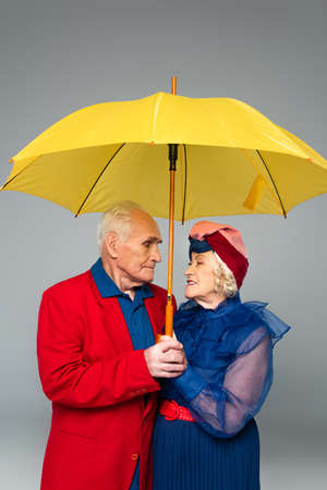 elderly man in red blazer and woman in blue dress and turban holding yellow umbrella isolated on grayの写真素材