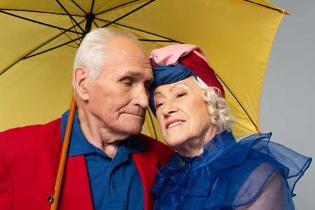 elderly man with yellow umbrella hugging wife in blue dress and turban isolated on gray