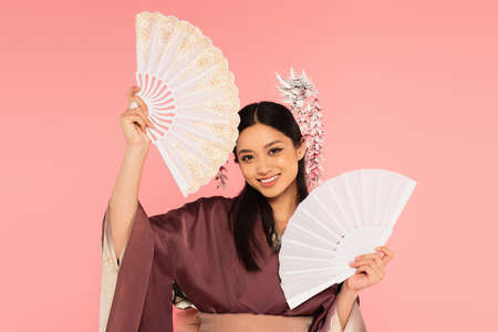 Smiling asian woman holding fans isolated on pink