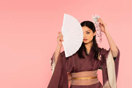 Asian woman with traditional kanzashi in hair covering face with fan isolated on pinkの写真素材