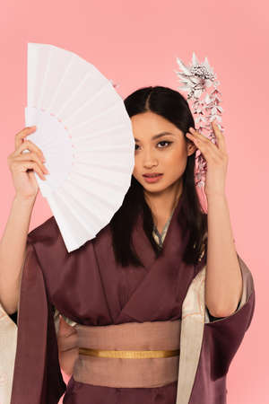 Asian woman in traditional clothes holding fan isolated on pink