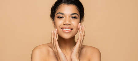 happy african american woman with bare shoulders touching clean face isolated on beige, bannerの写真素材