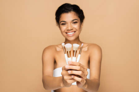 blurred and cheerful african american woman with bare shoulders holding cosmetic brushes isolated on beigeの写真素材