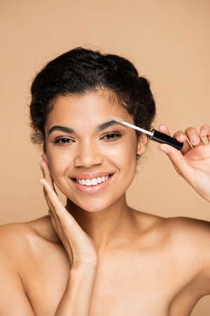 happy african american woman with bare shoulders styling eyebrow isolated on beige