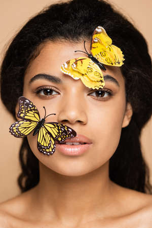 young african american woman with decorative butterflies on face looking at camera isolated on beigeの写真素材