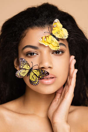 brunette african american woman with decorative butterflies on face looking at camera isolated on beigeの写真素材