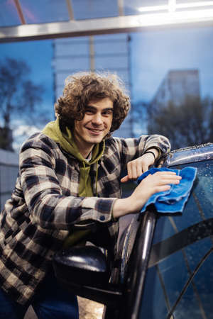 Curly man smiling at camera while cleaning wet car outdoors in eveningの写真素材