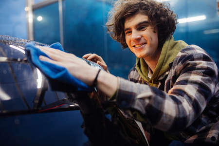 Smiling man in casual clothes cleaning car with rag on blurred foreground