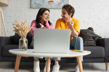 smiling young couple sitting on couch, looking at each other and pointing with fingers at laptop in...