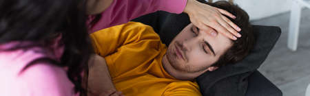 young woman measuring temperature with palm on forehead of sick young man lying on couch with closed eyes at home, bannerの写真素材