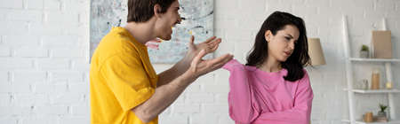 angry young man yelling with outstretched hands near girlfriend in living room, bannerの写真素材