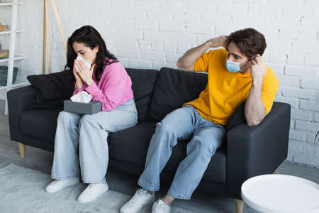 diseased young woman sitting on couch and blowing nose with paper napkin near boyfriend in medical...