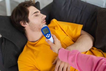 female hand with non-contact pyrometer near diseased young man lying on couch with closed eyes at homeの写真素材