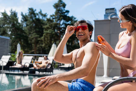 Smiling woman in swimsuit holding sunscreen near friend and swimming pool