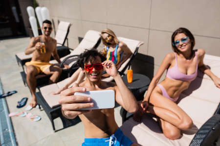 Smartphone in hand of man taking selfie near friends in swimwear on deck chairs