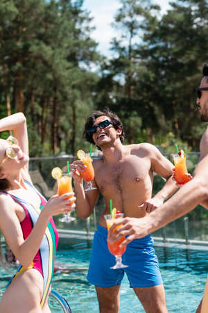 Happy man with cocktail standing near multiethnic friends and swimming pool during weekend