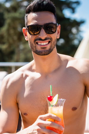 Smiling shirtless muslim man in sunglasses holding cocktail outdoorsの写真素材