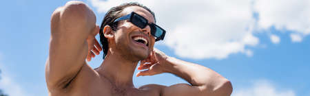 low angle view of young shirtless man in sunglasses smiling against blue sky, bannerの写真素材