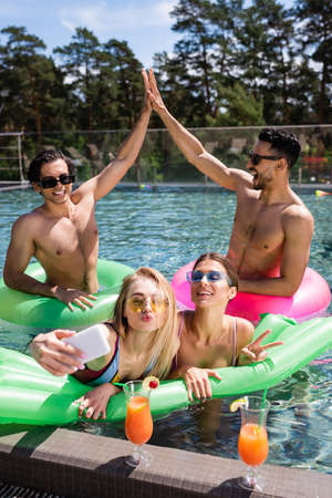 happy interracial men giving high five near women taking selfie in swimming poolの写真素材