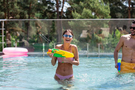 young woman with water pistol laughing in swimming pool near arabian man