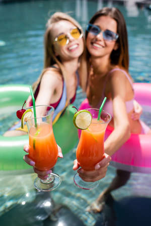 selective focus of cocktails glasses in hands of pretty women in swimming pool