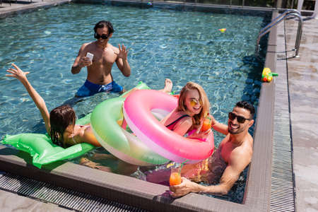 happy man with smartphone waving hand near interracial friends resting in swimming pool