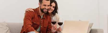 Smiling woman hugging boyfriend with glass of wine near laptop, bannerの写真素材
