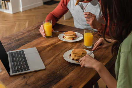 Cropped view of couple having breakfast with pancakes and orange juice near laptop