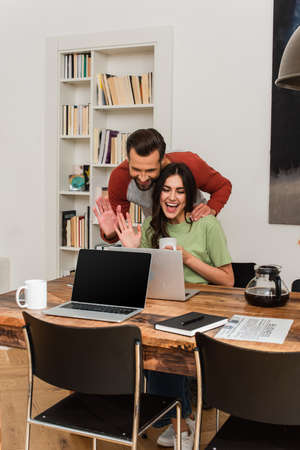 Cheerful couple having video call on laptop near coffee and newspaperの写真素材