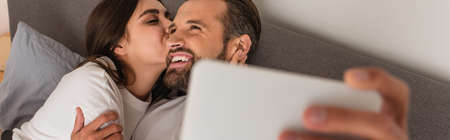 Woman kissing cheerful boyfriend during selfie on blurred smartphone, bannerの写真素材