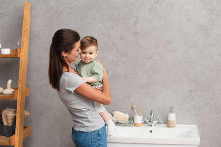 Cheerful mother embracing toddler son near sink in bathroom
