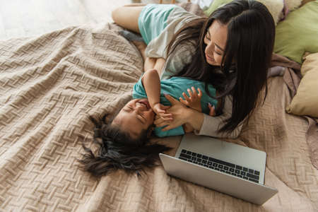 Overhead view of mother having fun with asian child near laptop on bedの写真素材