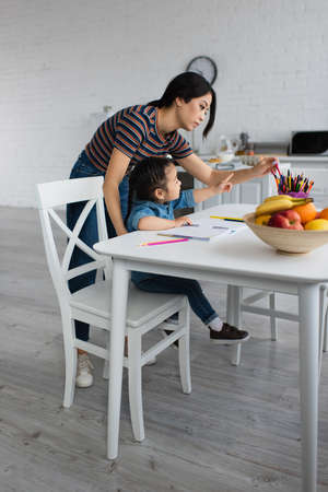 Side view of asian kid pointing at color pencils near mother and fruitsの写真素材