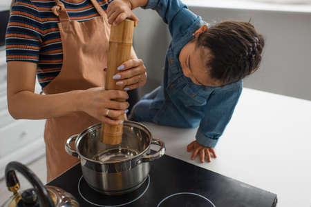 Asian kid holding salt mill while mother cooking in kitchenの写真素材