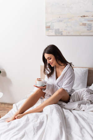 joyful young woman holding cup of coffee and saucer in bedroomの写真素材
