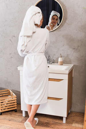 happy young woman applying face cream and looking at mirror in bathroom