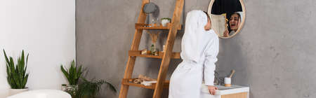 young woman in bathrobe with towel on head brushing teeth in bathroom near mirror, banner