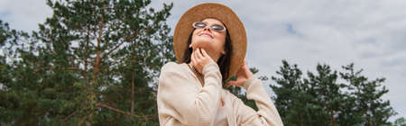 low angle view of cheerful young woman in sunglasses and straw hat near forest, banner