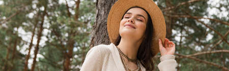 cheerful young woman adjusting straw hat and smiling in woods, banner