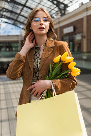 young woman in sunglasses holding yellow tulips and shopping bagsの写真素材