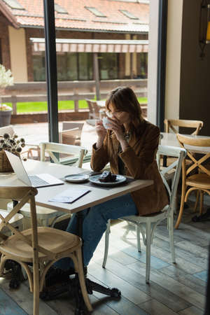 young woman in eyeglasses looking at laptop near chocolate cake and drinking coffee in cafeの写真素材
