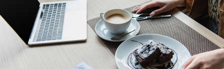 Cropped view of woman sitting near coffee, dessert and laptop with blank screen in cafe, banner