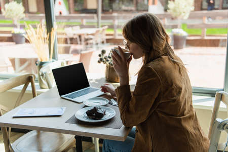Side view of young woman drinking coffee near dessert and laptop in cafeの写真素材