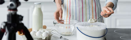 partial view of woman in apron holding spoon with flour near blurred digital camera, banner