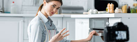 pretty culinary blogger showing cooking time near oven and blurred digital camera, banner