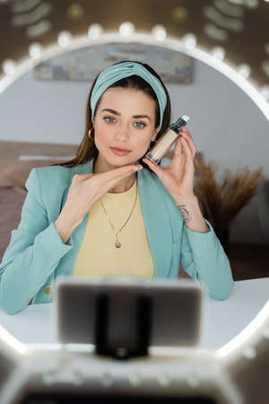 blurred phone holder with circle lamp near beauty blogger showing face foundation
