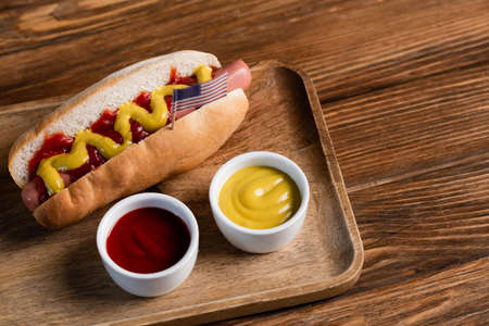 hot dog with small usa flag near bowls with sauces on wooden tableの写真素材
