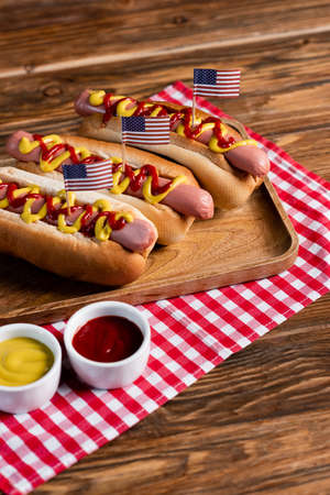 hot dogs with small usa flags on tray and plaid napkin near ketchup and mustard on wooden tableの写真素材