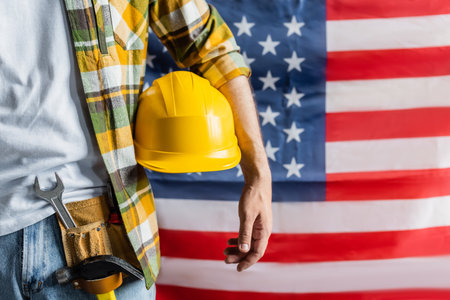 cropped view of workman in plaid shirt and tool belt holding hardhat near blurred usa flag, labor...