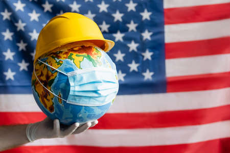 cropped view of man in latex glove holding globe with hardhat and medical mask near blurred usa flag, labor day conceptの写真素材